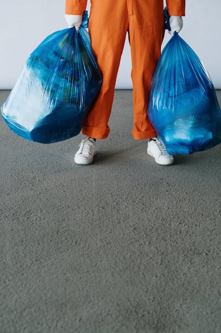 A person dressed in orange work overalls and white footwear is standing on a smooth, grey concrete floor, holding two large transparent blue plastic rubbish bags filled with waste material, one in eac