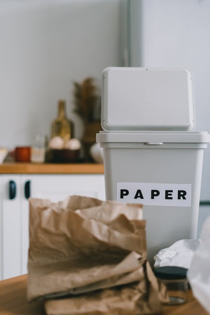 A recycling bin made of light grey plastic with a hinged lid, situated on a work surface or table in a domestic kitchen or utility area, with the word 'PAPER' printed in bold black letters on a white label affixed to the front of the bin. In the foreground, a crumpled brown paper bag is placed on the same surface, with some scattered white paper or tissue nearby. The background shows blurred kitchen elements, including a countertop with various bottles and containers, possibly for food or household supplies, and a section of a cabinet or wall. The lighting is natural and evenly diffused, highlighting the textures of the paper bag and the smooth surface of the bin. This scene aligns with concepts of household waste segregation and independent rubbish disposal, supporting the context of rubbish removal services that facilitate on-site waste sorting and collection.